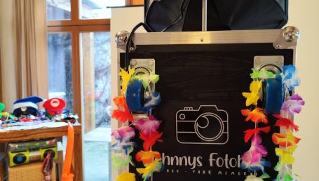 Kamerafotobox mit buntem Blumenkranz und Rollen, im Hintergrund Spielzeug.